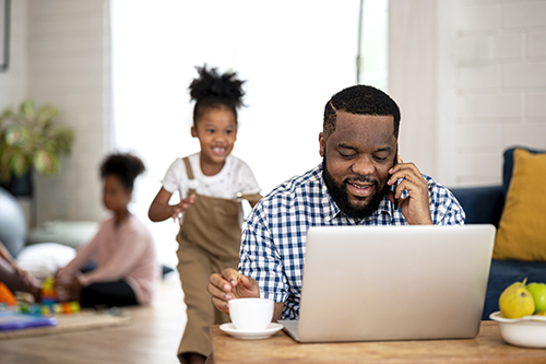 A African father working form home talking on mobile phone, while daughter running from behind being to sill covering his eyes to stop him from working.